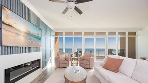 A spacious living room with a large TV, comfortable seating, a ceiling fan, and a balcony with a view.