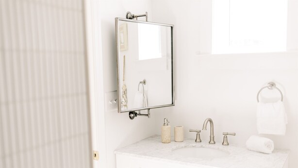 A bathroom with a white vanity, a mirror, a sink with two faucets, a towel rack, and a toilet paper holder.