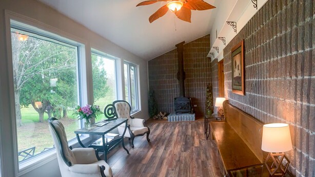 A spacious room with a wooden floor, a dining table with chairs, a fireplace, and large windows offering a view of the outdoors. - Beautiful farmhouse with horses, pet friendly & close to Prince Edward County. (Brighton)