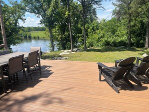 Terraza de madera con mobiliario exterior, vista a un lago y vegetación circundante.