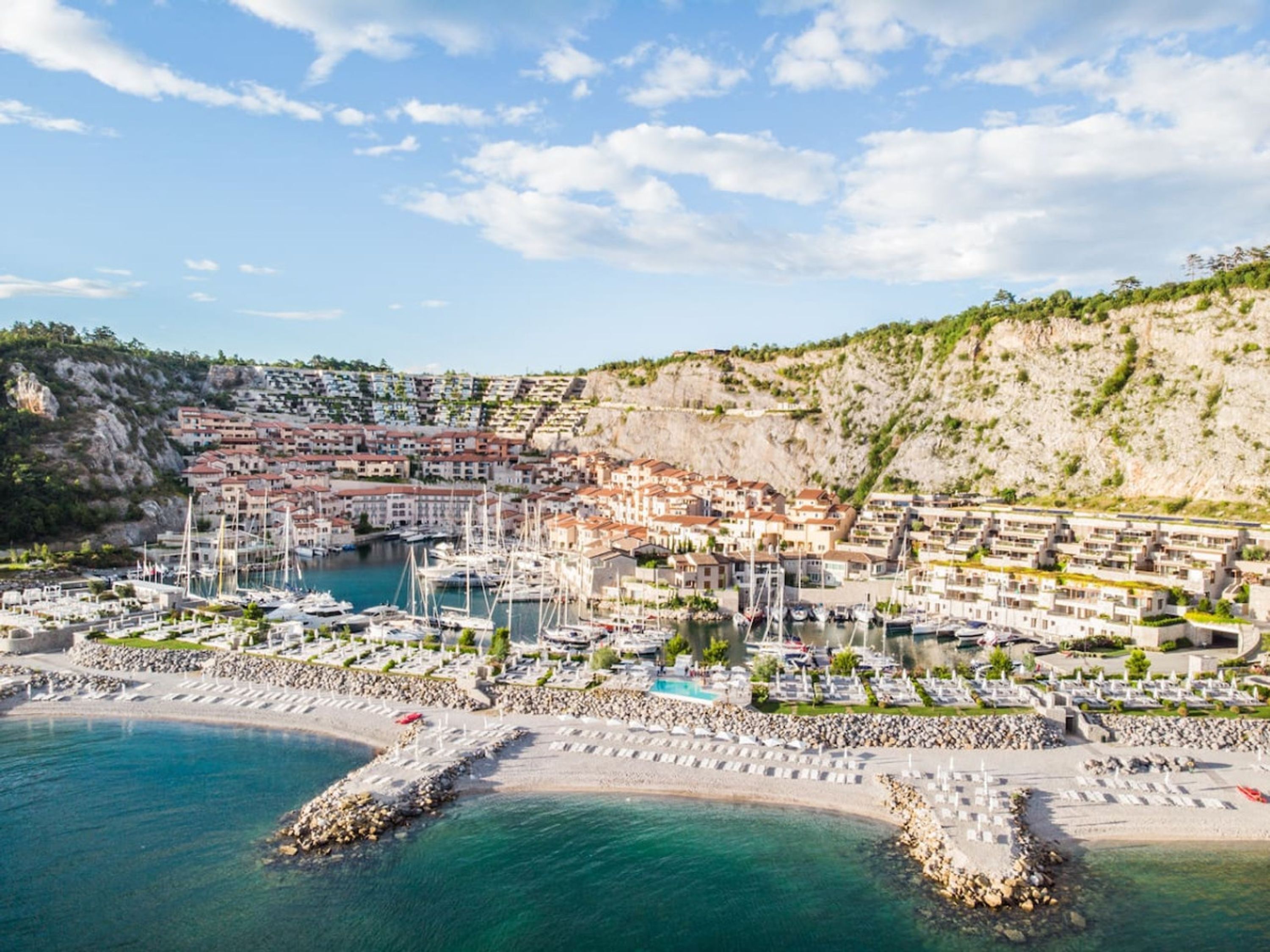 Una località costiera con un porto turistico, spiaggia e edifici arroccati su una collina.
