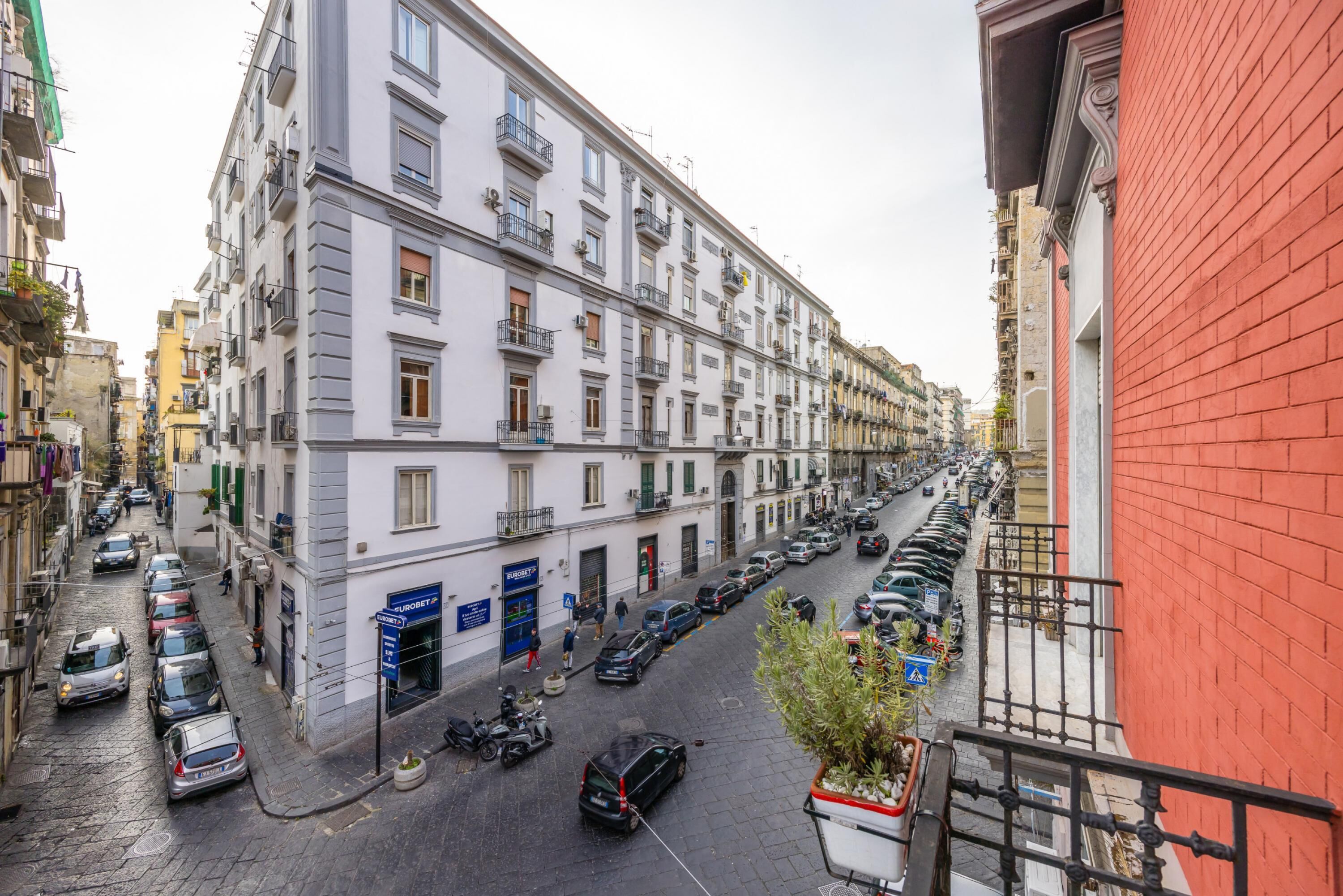 A narrow street lined with multi-story buildings, parked cars, and a few pedestrians.