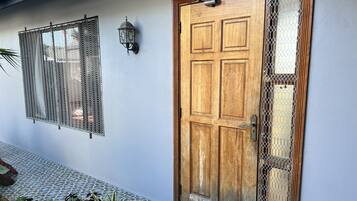 A wooden door with a metal mesh window, a wall-mounted light, and a tiled floor with plants.