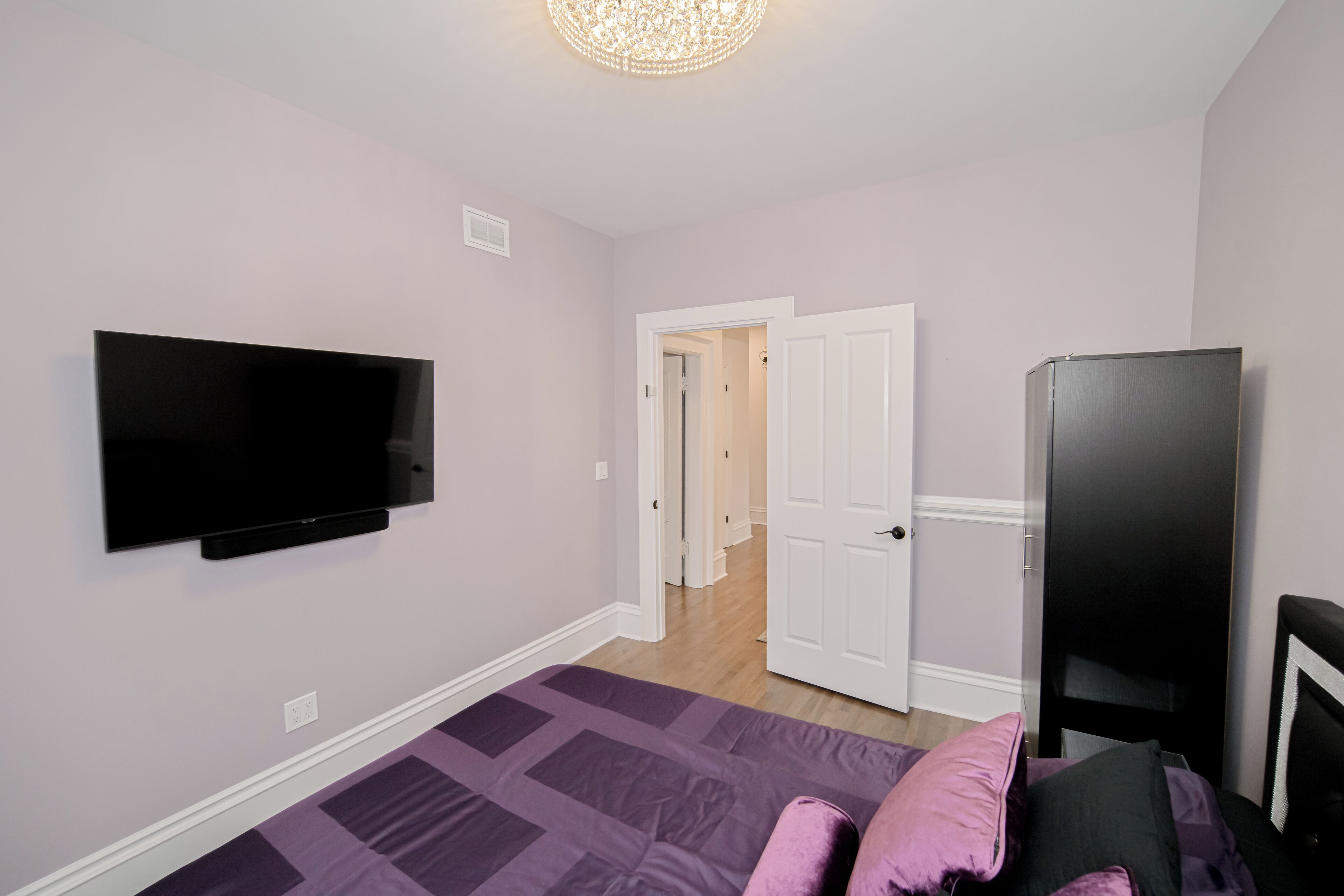 A bedroom with a flat-screen TV mounted on the wall, a bed with a purple quilt, and a black wardrobe.