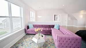 A spacious living room with a large window, a purple sectional sofa, a glass coffee table, and a colorful patterned rug.
