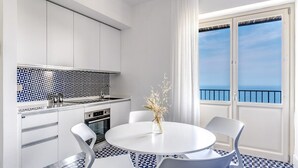 A modern kitchen with white cabinets and a blue and white patterned backsplash. There is a dining table with four chairs and a vase with dried plants. A large window with a view of the sea is visible. - Le Arcate Giuliana by Feeling Italy (Massa Lubrense)