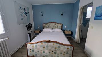 Quarto com cama, mesas de cabeceira, ventilador e janela.