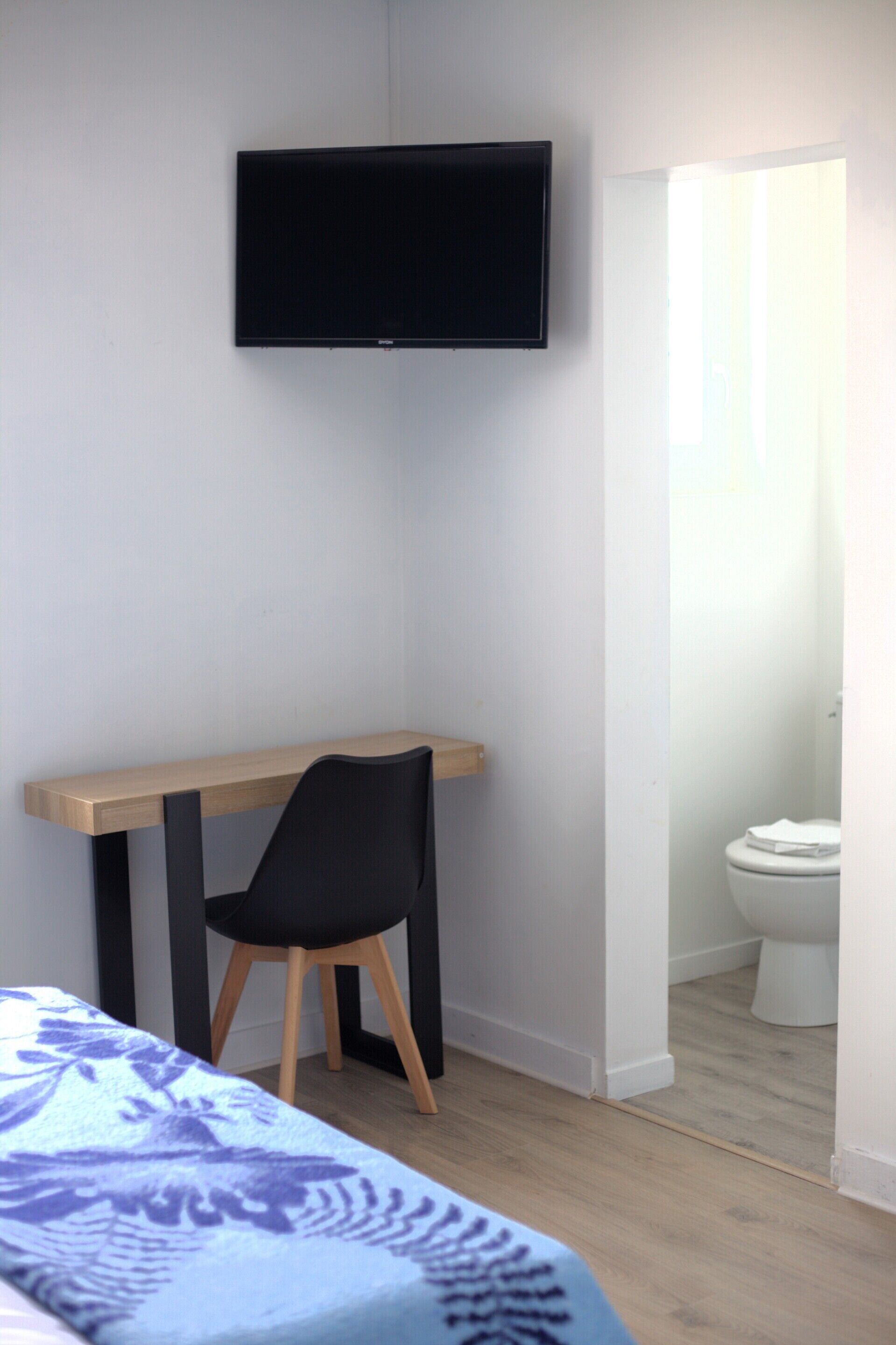 Ein Schlafzimmer mit einem Bett, einem Schreibtisch mit Stuhl, einem an der Wand befestigten Fernseher und einer Toilette, die durch eine offene Tür sichtbar ist.