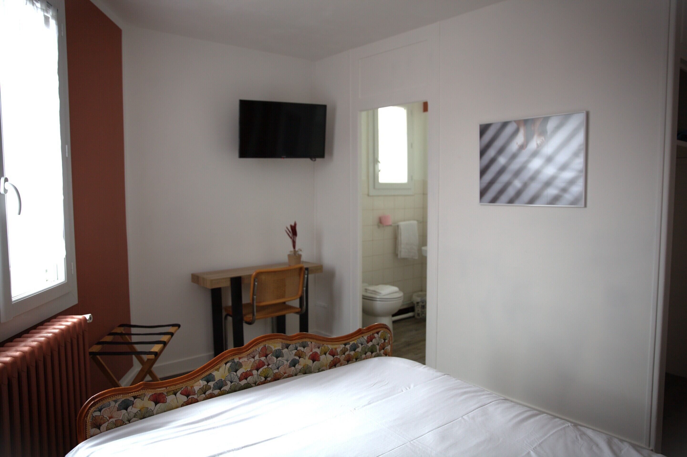 Ein Schlafzimmer mit einem Bett, einem an der Wand montierten Fernseher, einem kleinen Tisch mit Stuhl und einem Fenster.