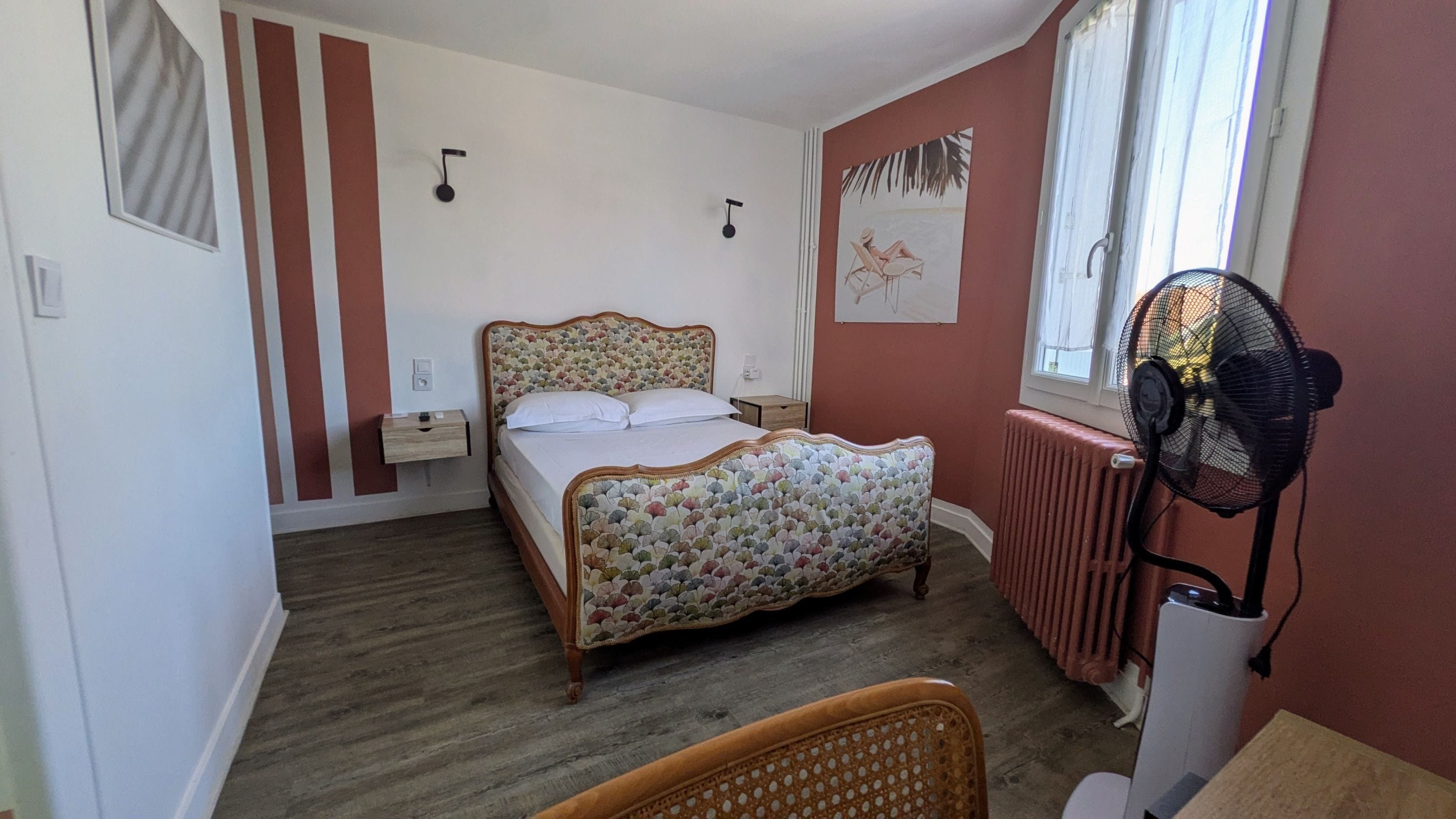 Ein Schlafzimmer mit Bett, Nachttischen, Heizkörper, Ventilator und Fenster.