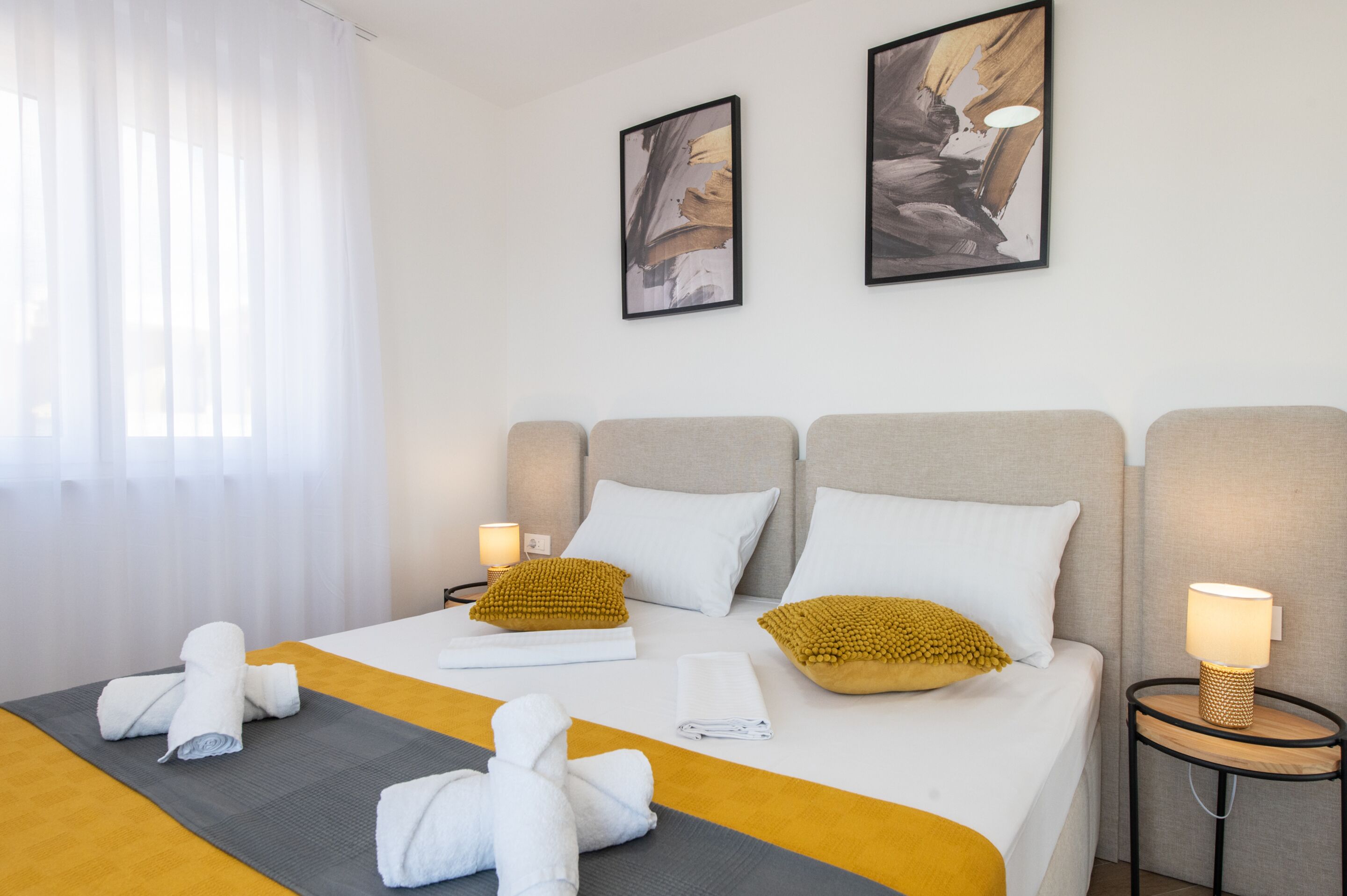 Hotellihuone, jossa on sänky, kaksi tyynyä, yöpöytälamppu, pyyheteline ja taideteos seinällä.