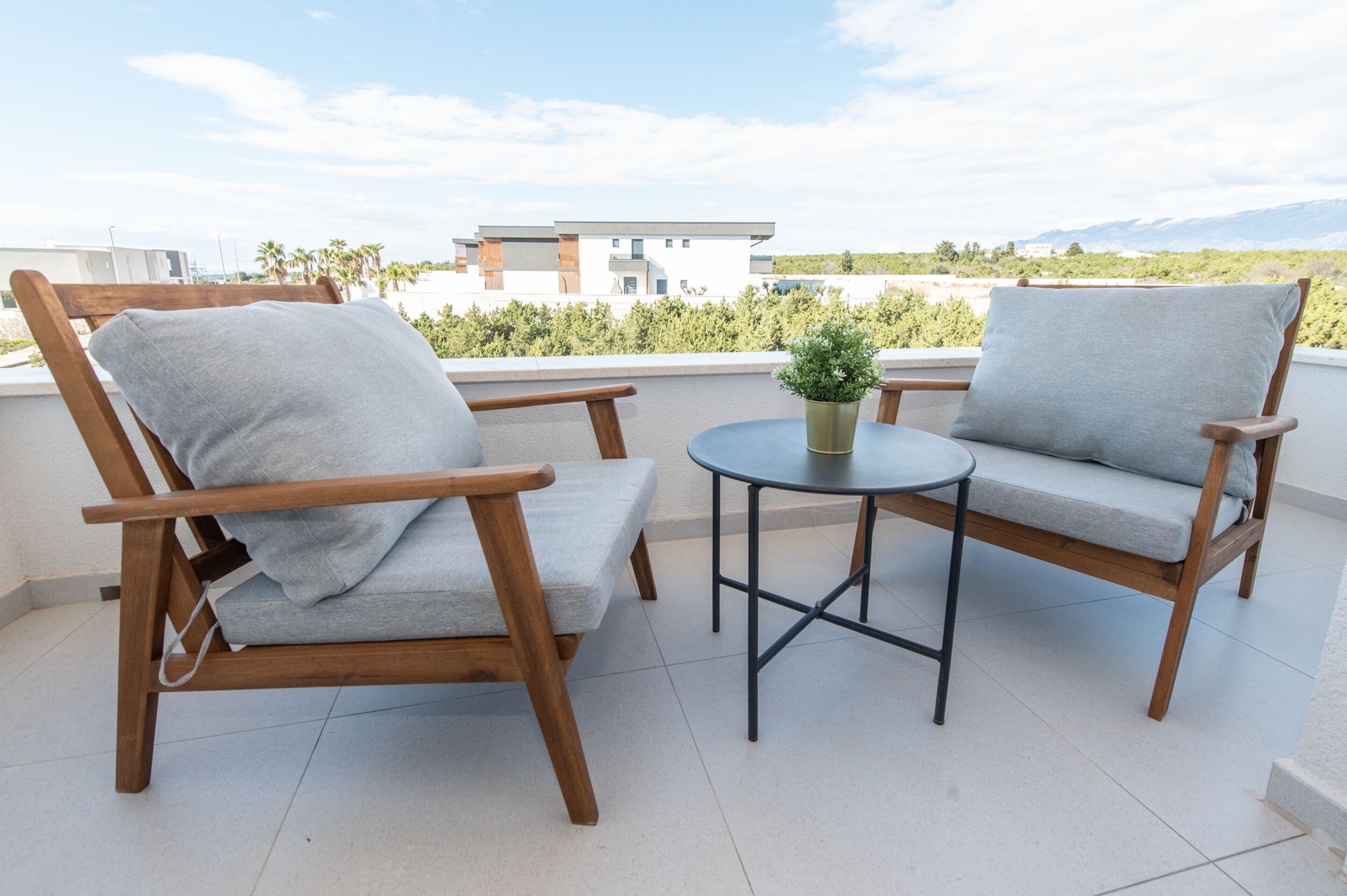 A balcony with two wooden chairs and a small round table, a potted plant, and a view of a modern building and distant mountains.