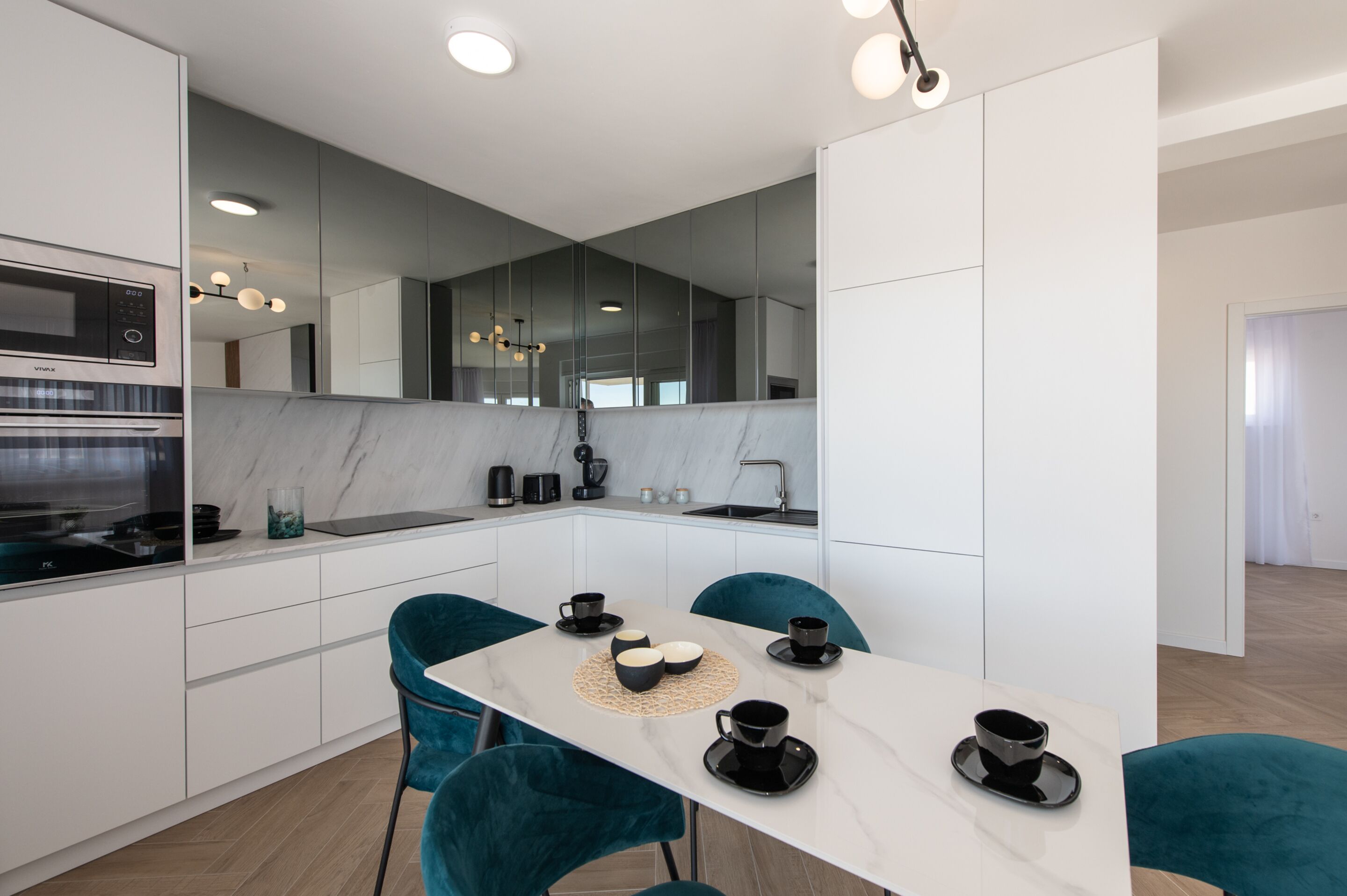 A modern kitchen with white cabinetry, a marble countertop, and a dining area with teal chairs and a white table.