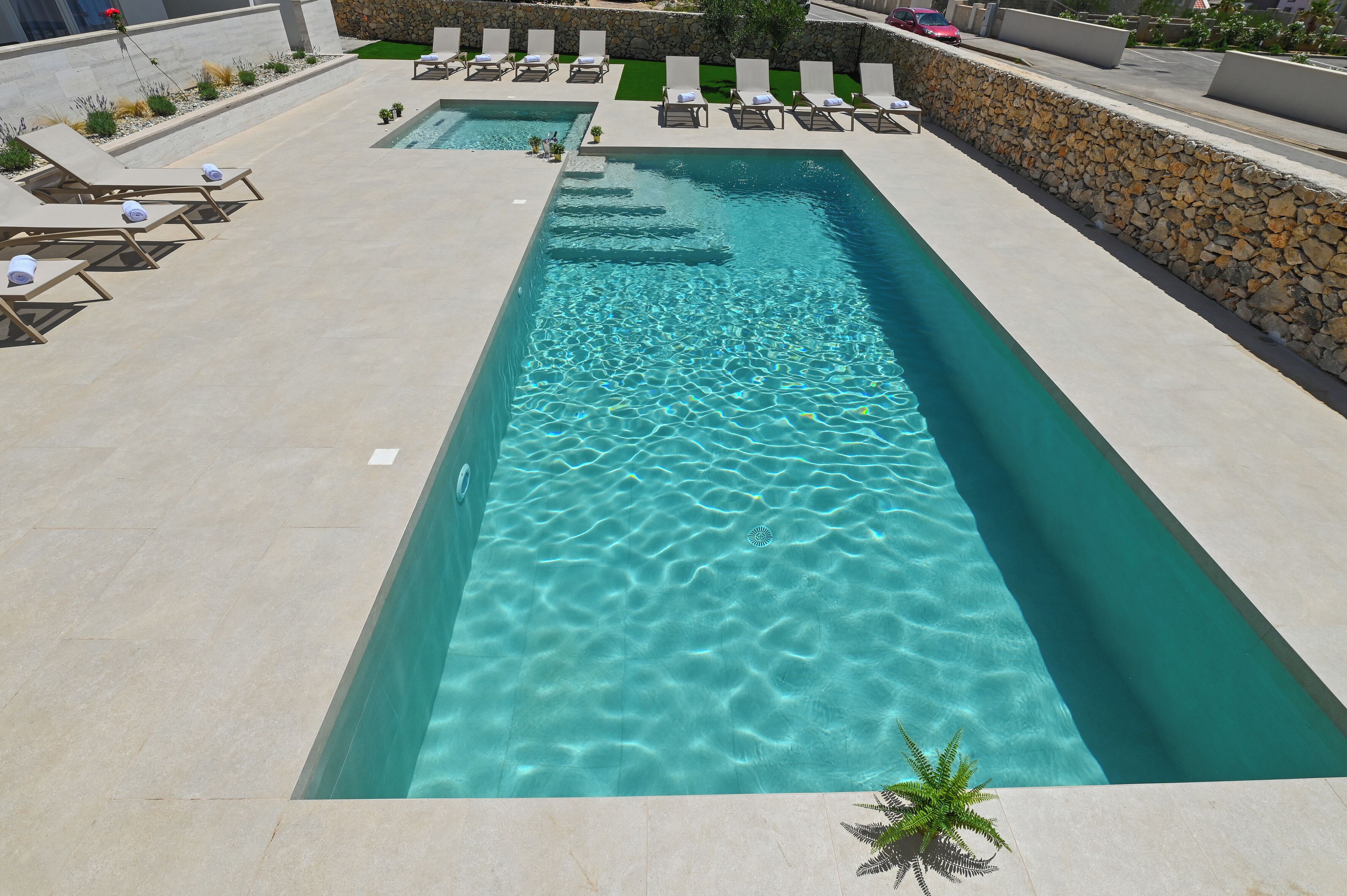A rectangular swimming pool with clear water, surrounded by lounge chairs and a stone wall.