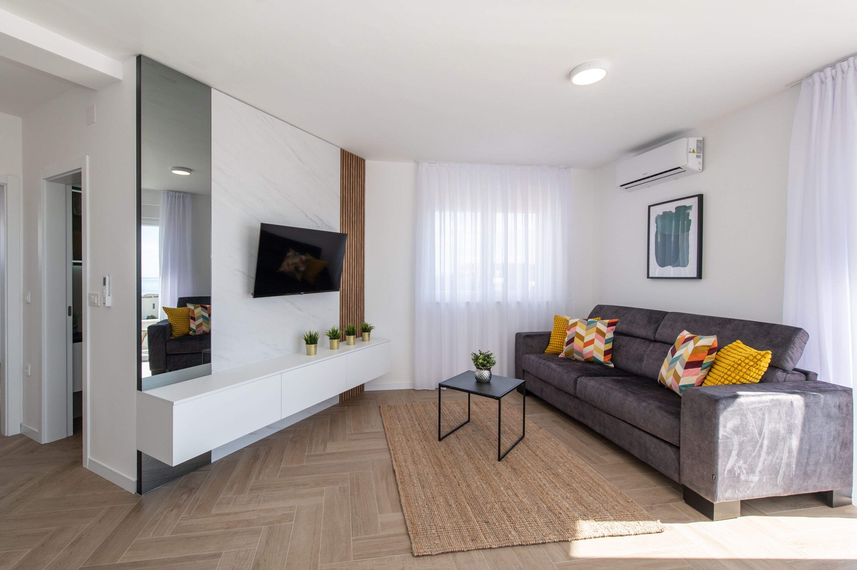 A modern living room with a grey sofa, a flat-screen TV mounted on a white wall, a small side table with a plant, and a large mirror.