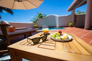 Une table en bois sur laquelle sont posés une paire de lunettes de soleil, une assiette de fruits et un verre de jus d’orange, le tout sur fond d’une piscine et d’un ciel bleu clair.