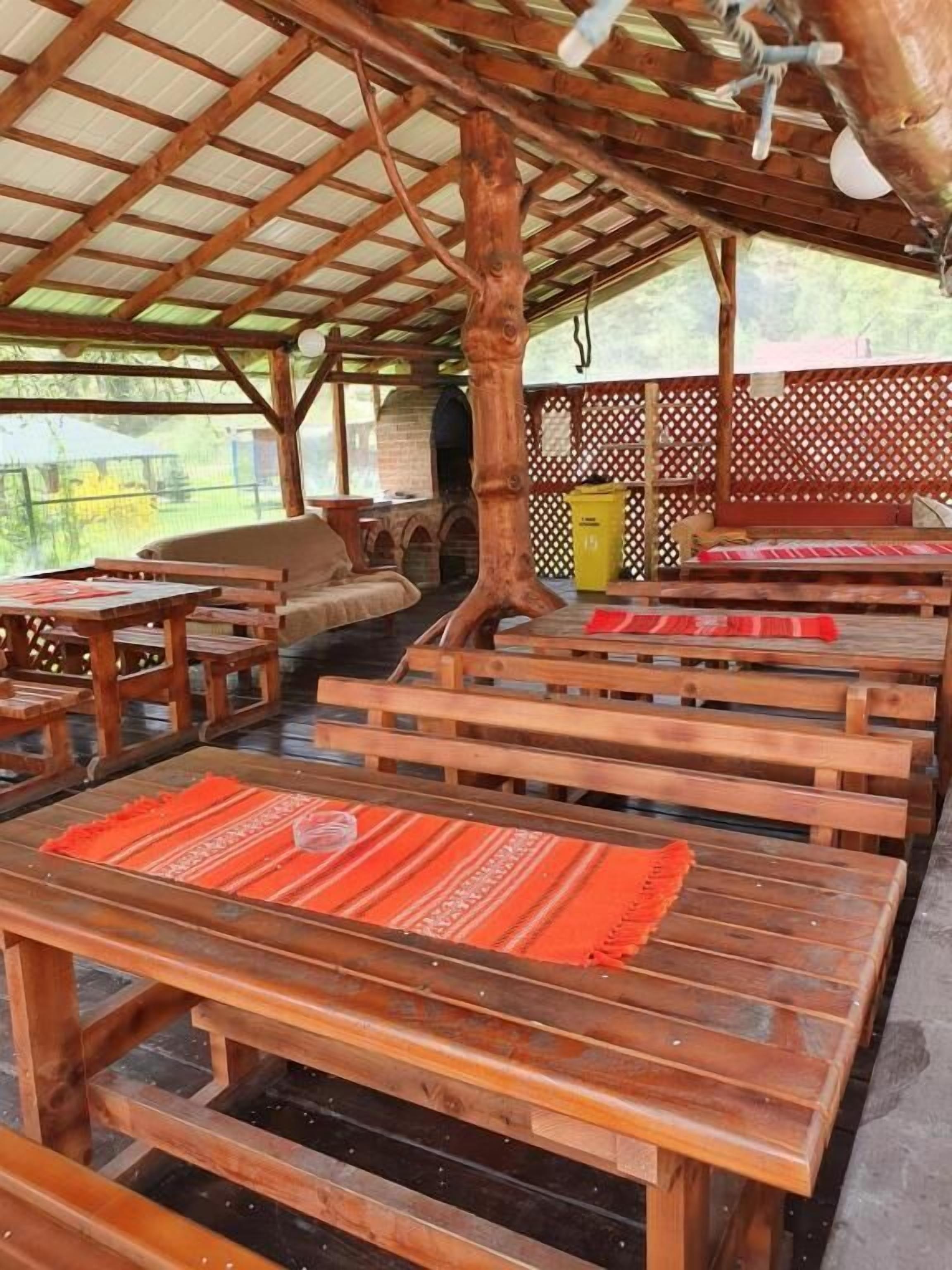 A covered outdoor seating area with wooden benches and tables, a wooden structure with a sloped roof, and a view of greenery in the background.