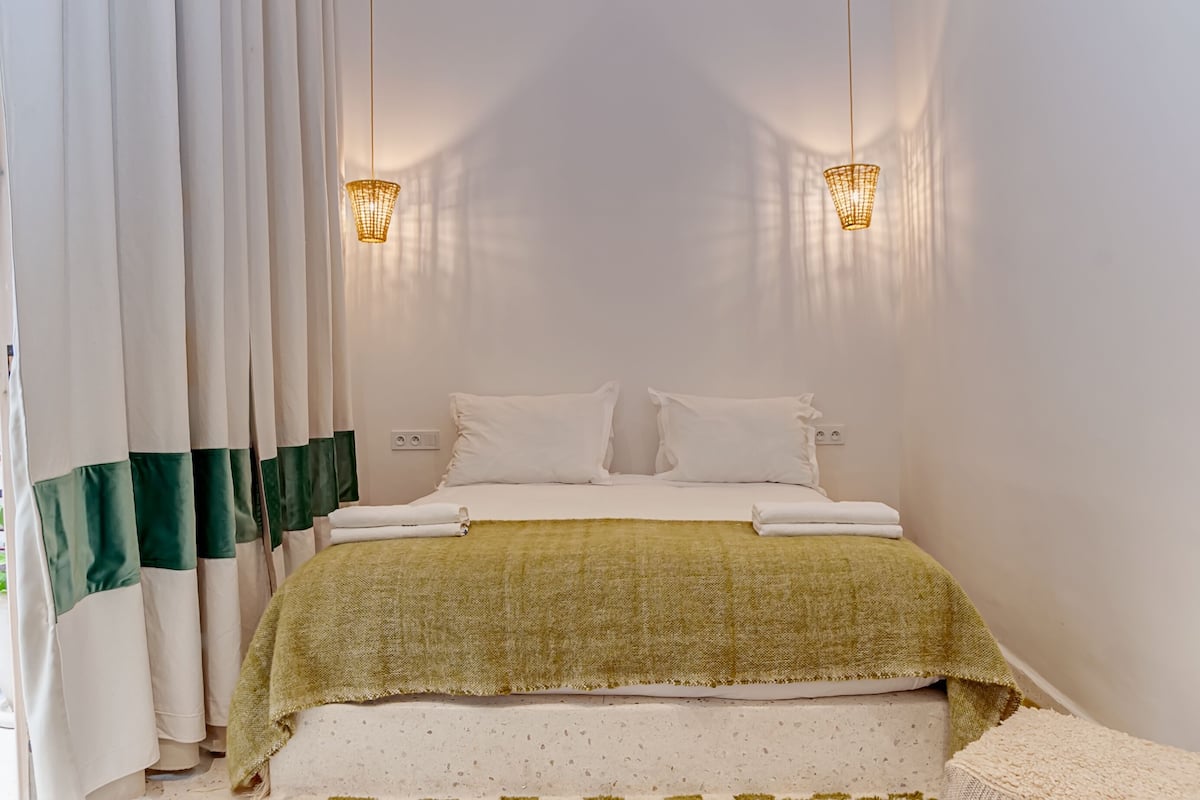 Un lit bien fait, avec du linge de lit blanc et un plaid vert, deux lampes de chevet et une fenêtre avec des rideaux.