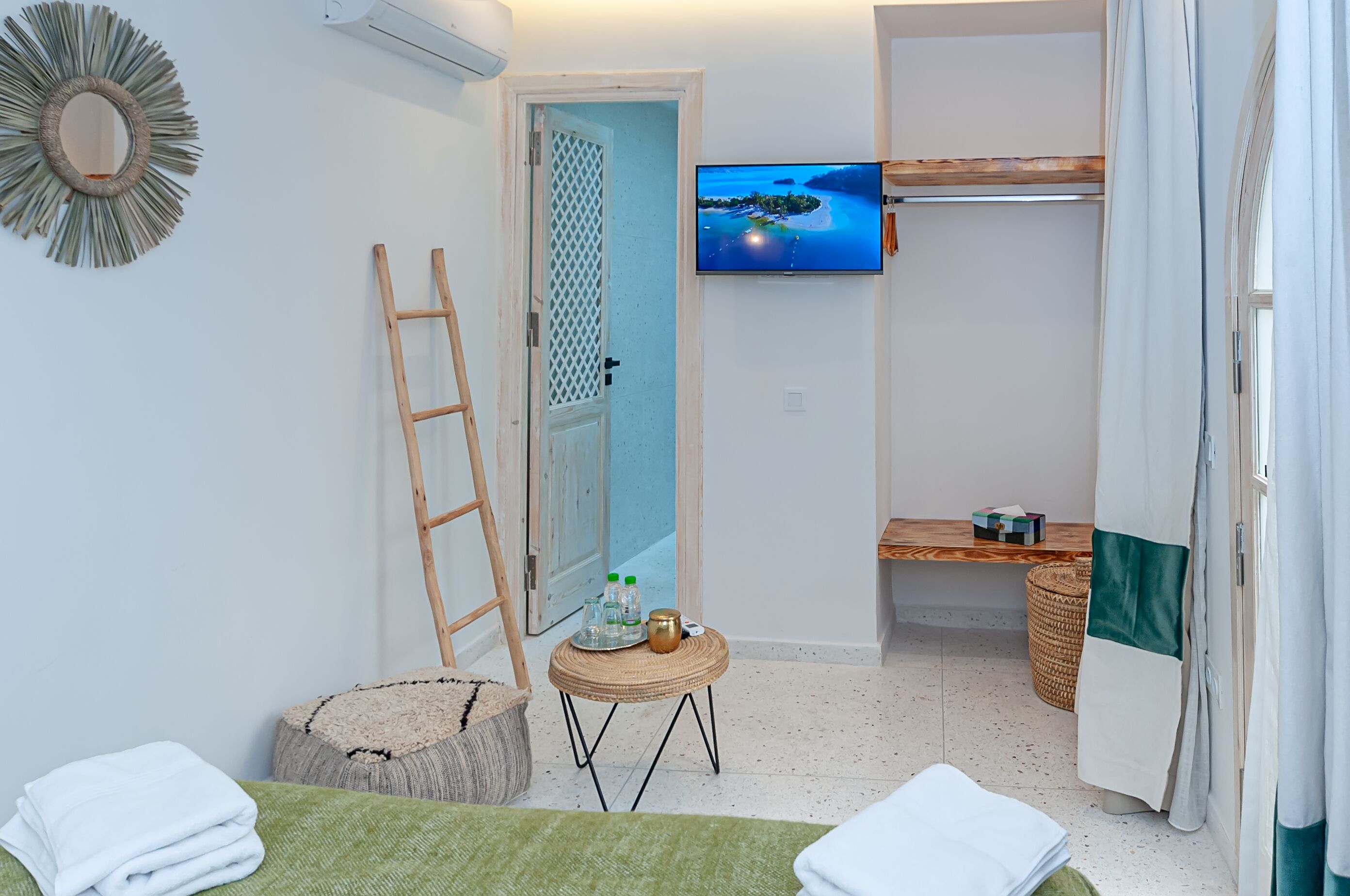 Moderni hotellihuone, jossa on seinälle asennettu televisio, puinen tikapuu, pieni pyöreä pöytä ja kori sekä vihreä matto.