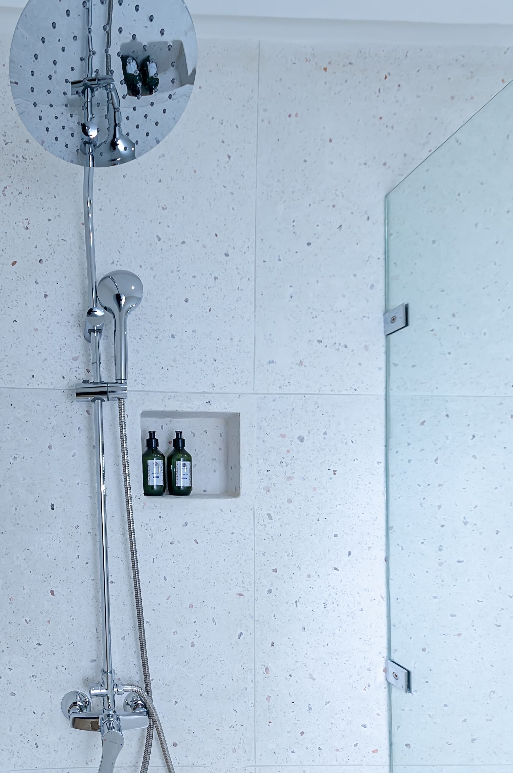 A modern shower with a handheld showerhead, a fixed showerhead, and a handheld shower hose. There is a small shelf with two bottles of shampoo and conditioner.