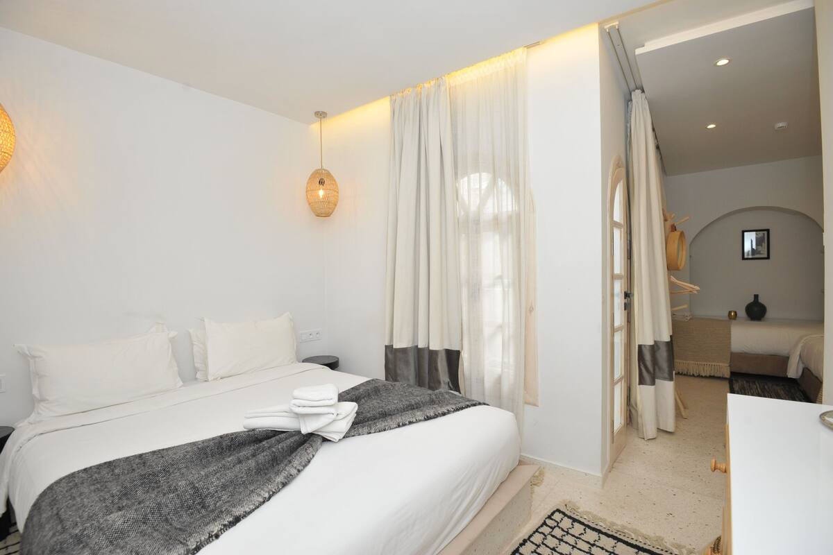 Une chambre d’hôtel moderne avec un lit soigneusement fait, un luminaire suspendu et une vue sur une autre chambre à travers une fenêtre avec des rideaux.