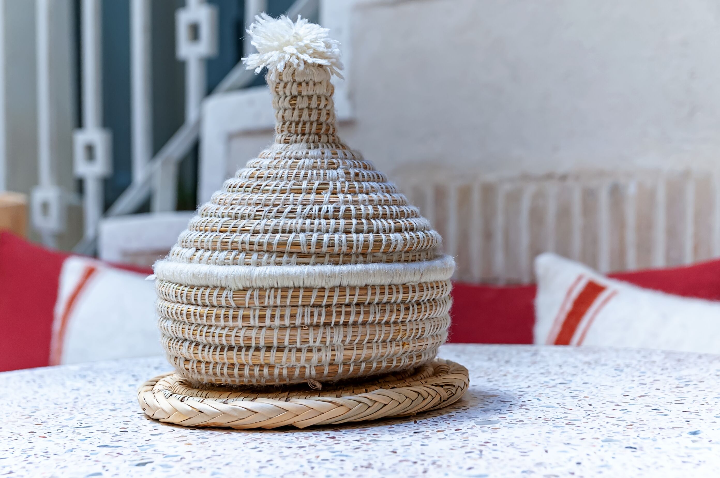 A woven basket with a tassel on top, placed on a woven coaster.
