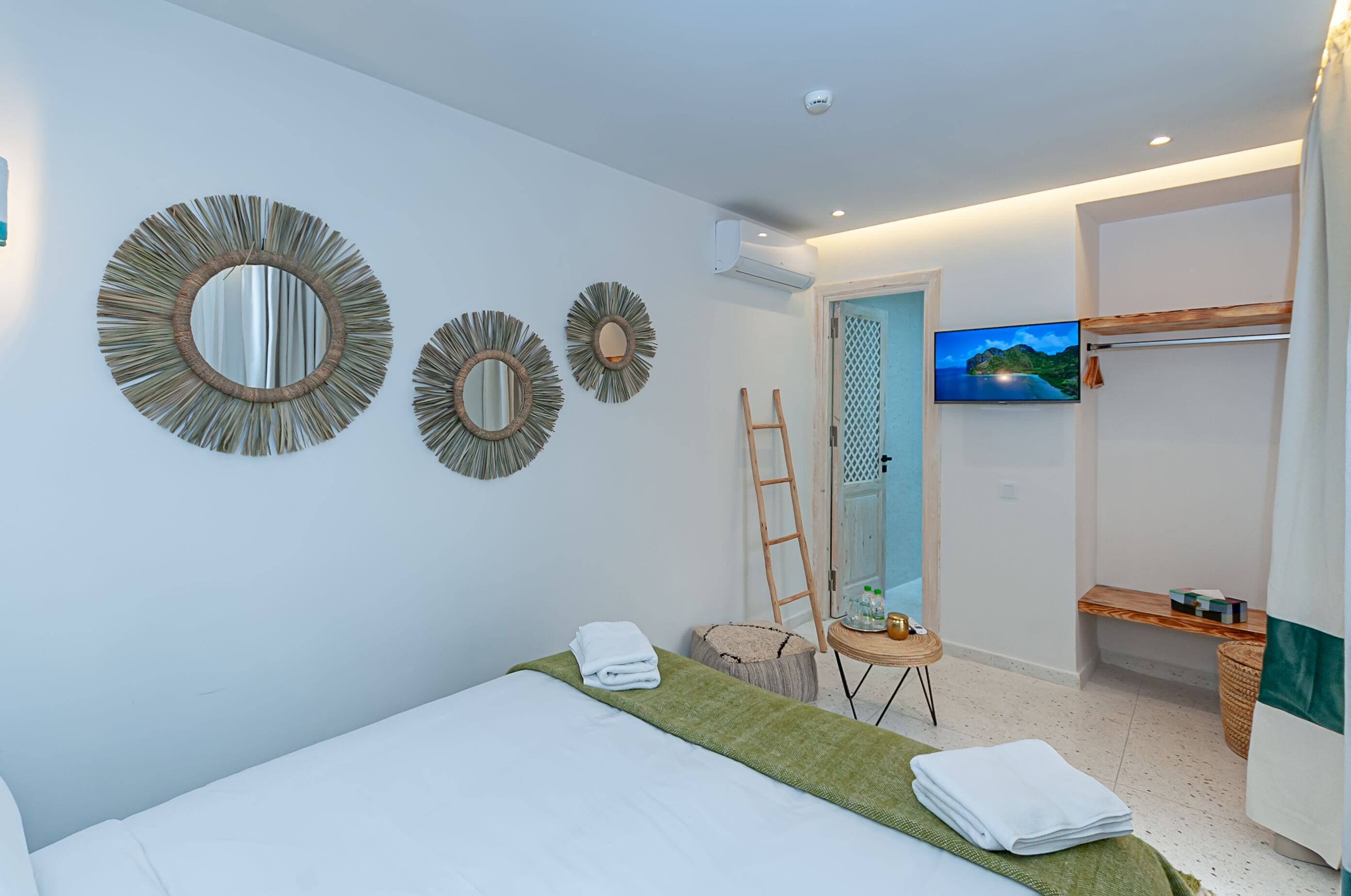 A modern hotel room with a bed, three wall mirrors, a TV, a wooden ladder, and a small table with toiletries.
