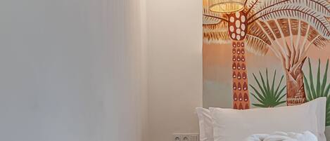 Une chambre avec un lit, un panier en osier, un luminaire suspendu et une fresque murale.