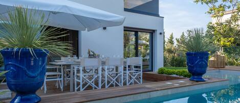 A modern outdoor dining area with a pool, white furniture, and large blue planters.
