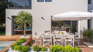 Ett modernt utomhusområde med plats för måltider, en pool, ett trädäck och vita möbler.
