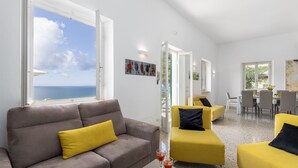 A modern living room with a grey sofa and yellow armchairs, a dining area with a table and chairs, and a view of the sea through large windows. - Villa Meraviglia 8, Emma Villas (Nisporto)
