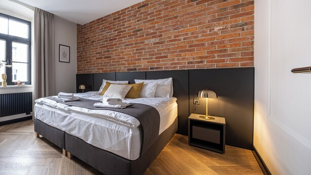 A modern bedroom with a brick wall, a bed with white bedding, a bedside table with a lamp, and a window with curtains. - Carvala Boutique Apartments (Czestochowa)