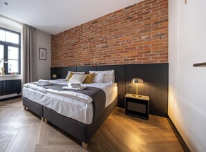 Un dormitorio moderno con pared de ladrillo, cama con ropa de cama blanca, mesita de noche con lámpara y ventana con cortinas.