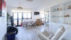 Un salon moderne avec un berceau blanc, une table à manger avec des chaises, un vélo d’appartement et des meubles en osier.