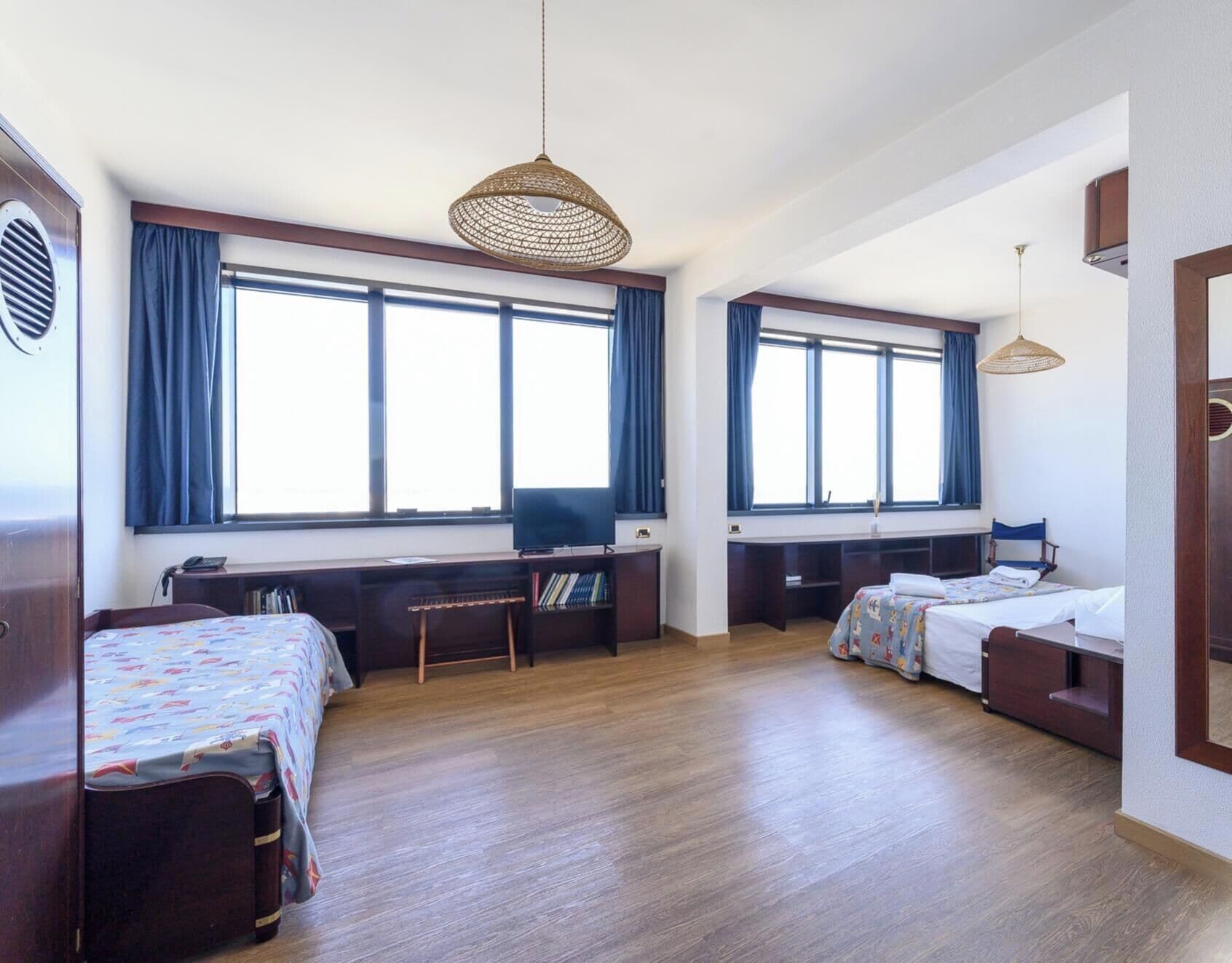 Une chambre d’hôtel avec deux lits, un bureau, une chaise, une bibliothèque et de grandes fenêtres avec des rideaux.