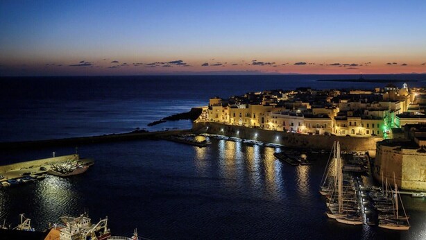 Une ville côtière au crépuscule, avec une structure fortifiée, un port rempli de bateaux et une vue dégagée sur la mer.