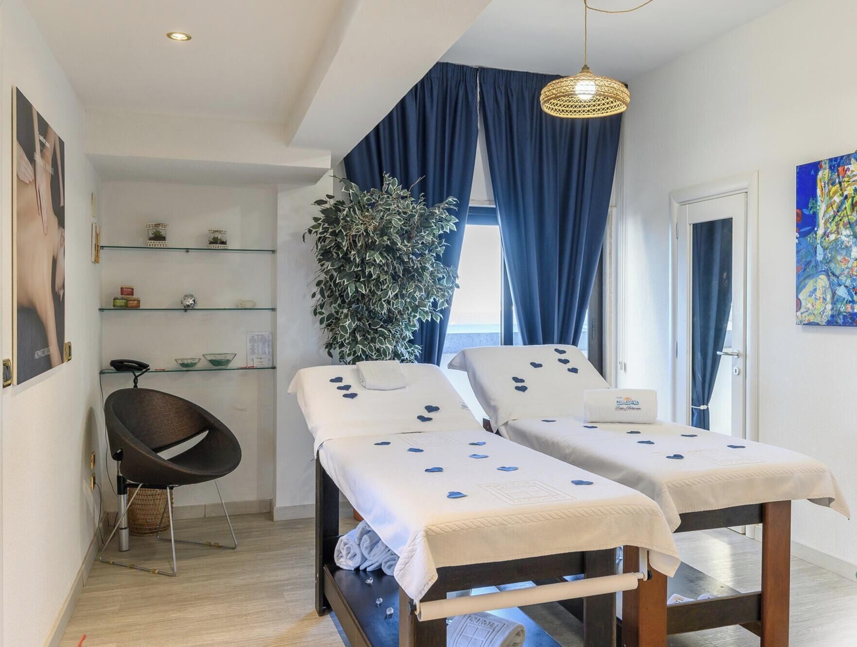 Una habitación con dos camas, una silla, un estante con objetos, una planta grande y un cuadro.