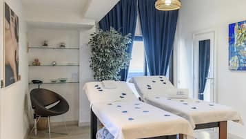 Una habitación con dos camas, una silla, un estante con objetos, una planta grande y un cuadro.