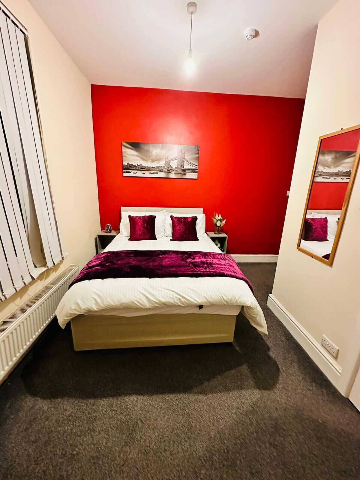 A bedroom with a red wall, a bed with white and purple bedding, a nightstand with a vase of flowers, and a picture of a bridge on the wall.