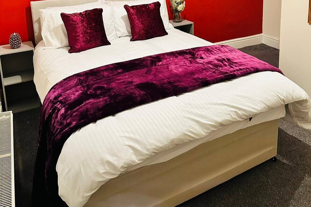 A hotel room with a red accent wall, a bed with white bedding and two red pillows, a black nightstand with a small decorative item, and a framed picture of a bridge.