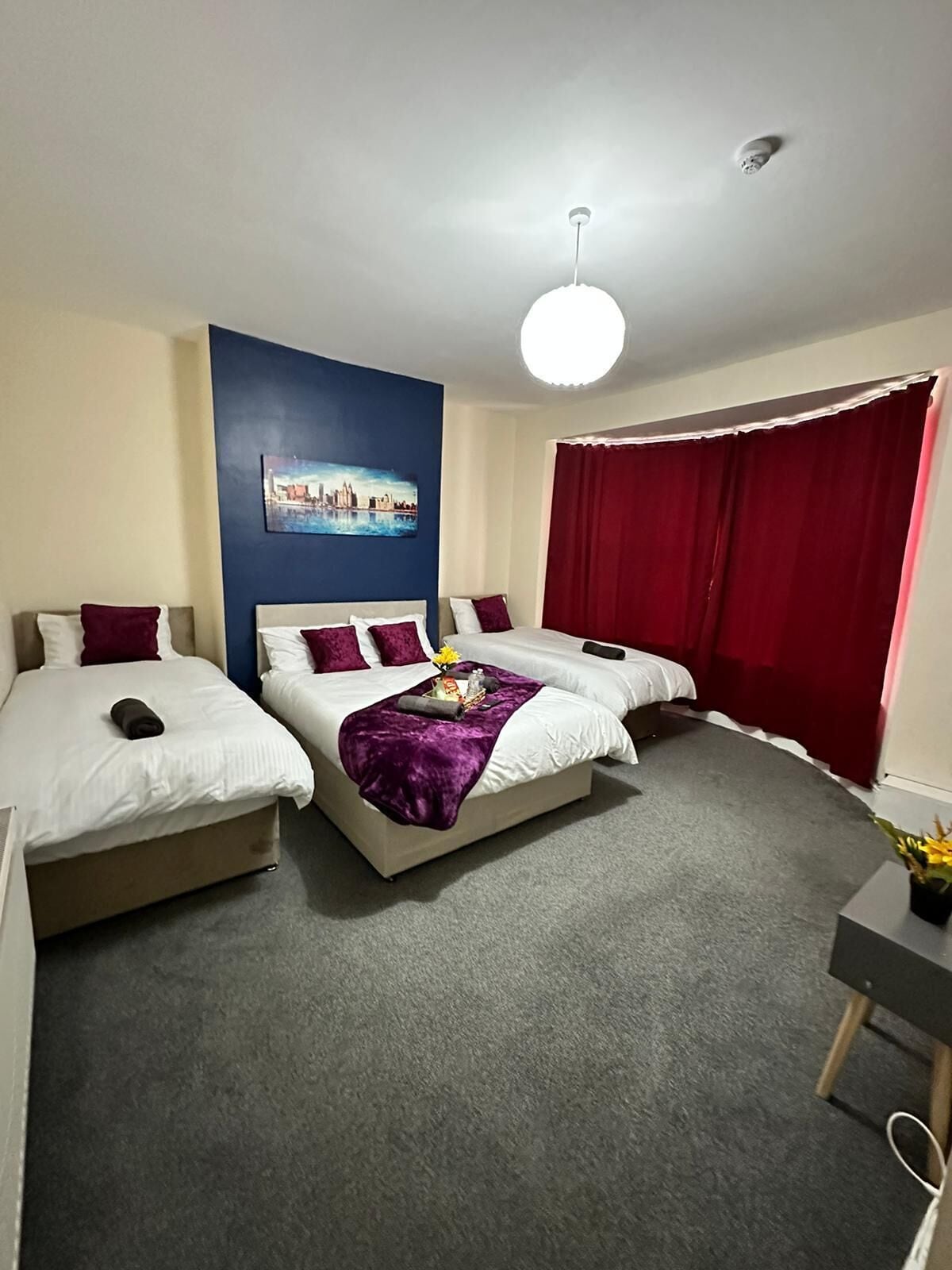 A hotel room with two single beds, a larger bed, a cityscape painting, a red curtain, and a ceiling light.