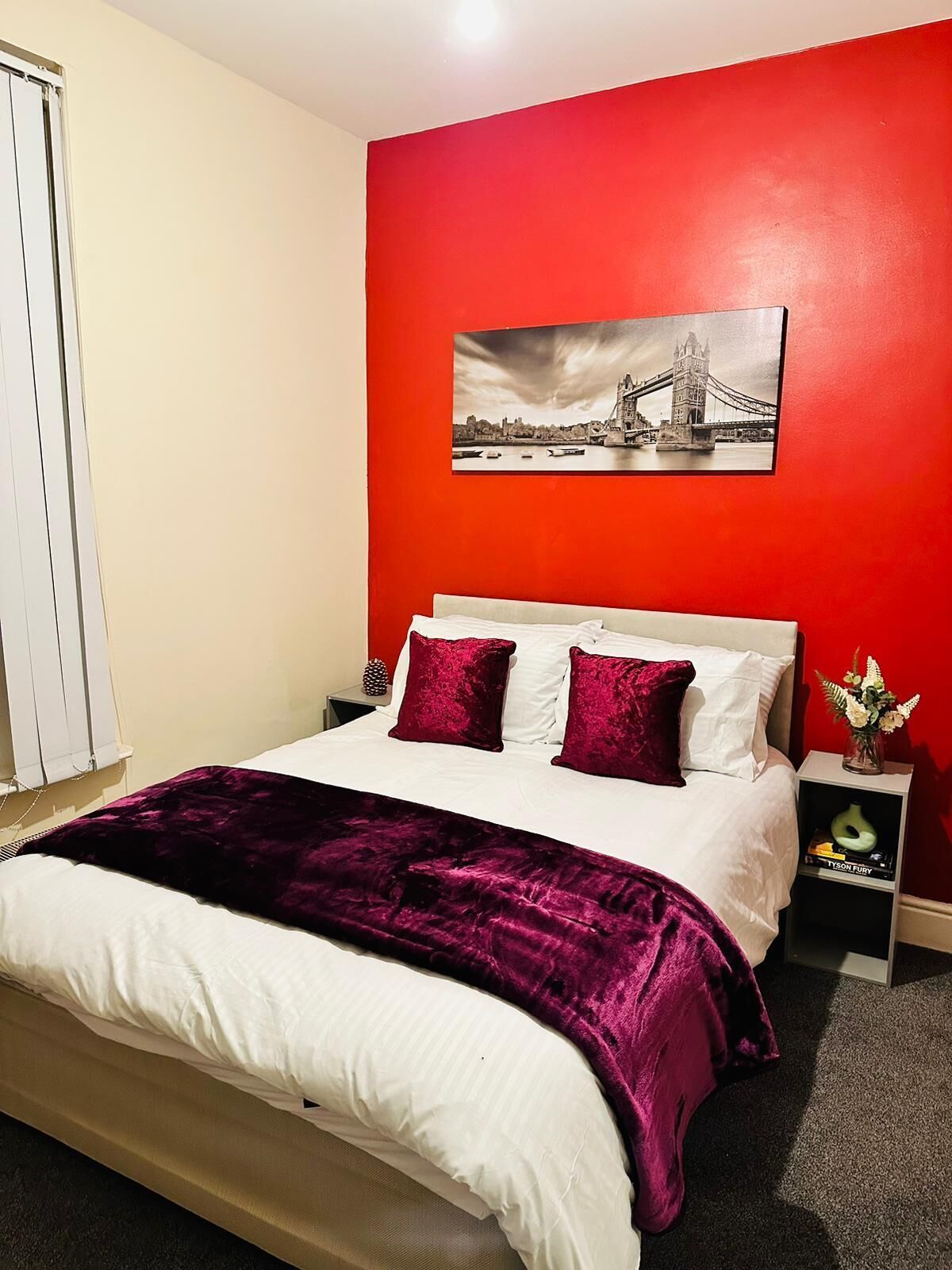 A bedroom with a red wall, a bed with white and purple bedding, a nightstand with a vase of flowers, and a picture of a bridge on the wall.