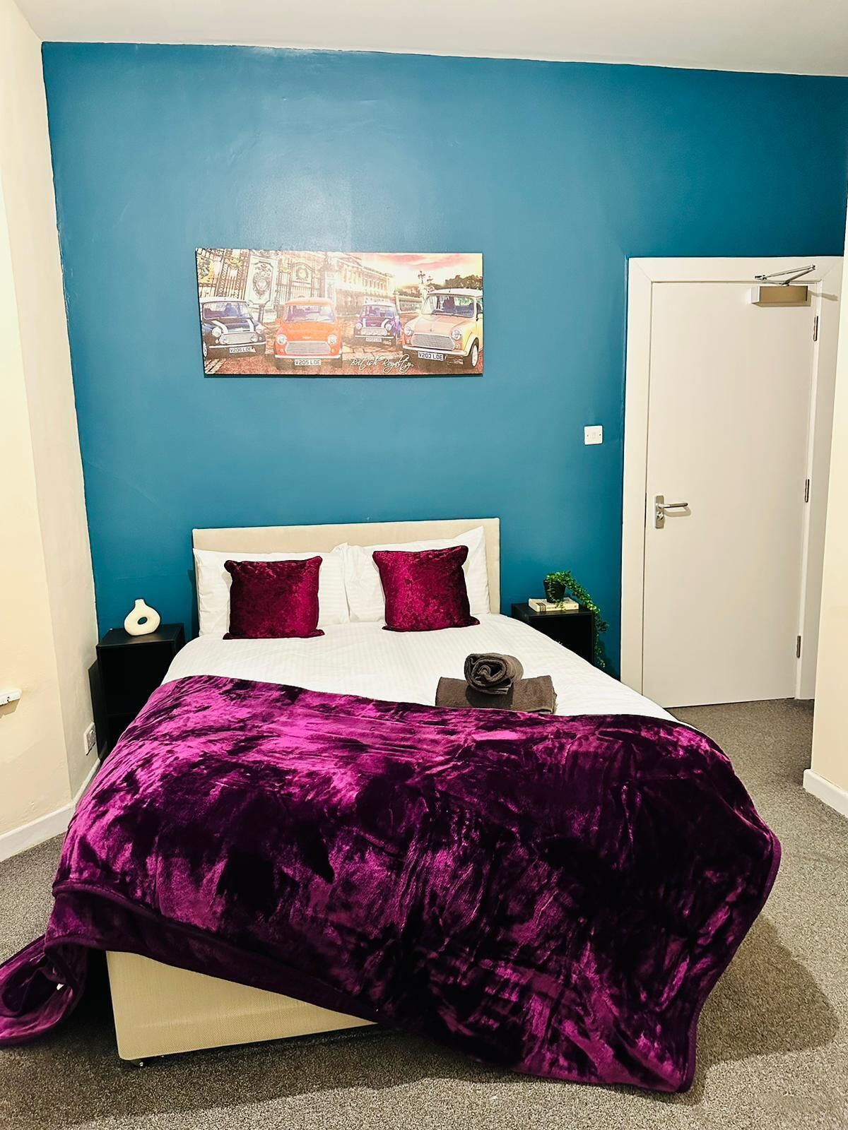 A bedroom with a blue wall, a bed with a purple blanket and red pillows, a nightstand with a lamp, and a door to the right.