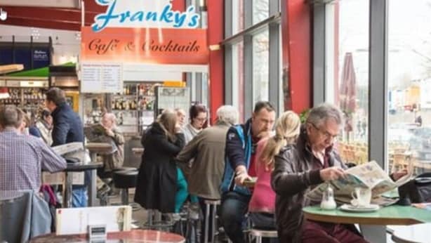 Un caffè vivace con tavoli, sedie, una bacheca dei menu e persone sedute o in piedi.