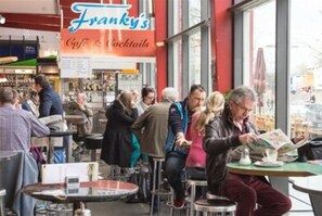 A busy café with tables and chairs, a menu board, and people sitting and standing. - Stadtflair-Apartment (Hannover)
