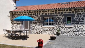 A stone building with a red-tiled roof, a patio with a table and chairs, and a blue umbrella.