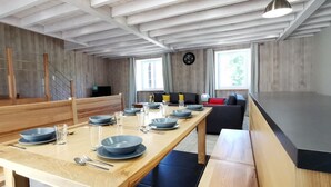 A modern dining area with a long wooden table set for six, a staircase, and a living area with a sofa and a TV.