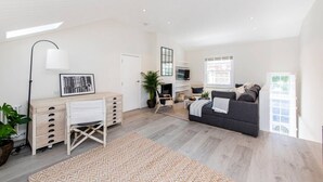 A spacious living area with a wooden desk, a dark sofa, and a large window providing natural light. - Divine 3-bed Mews House Battersea (London)