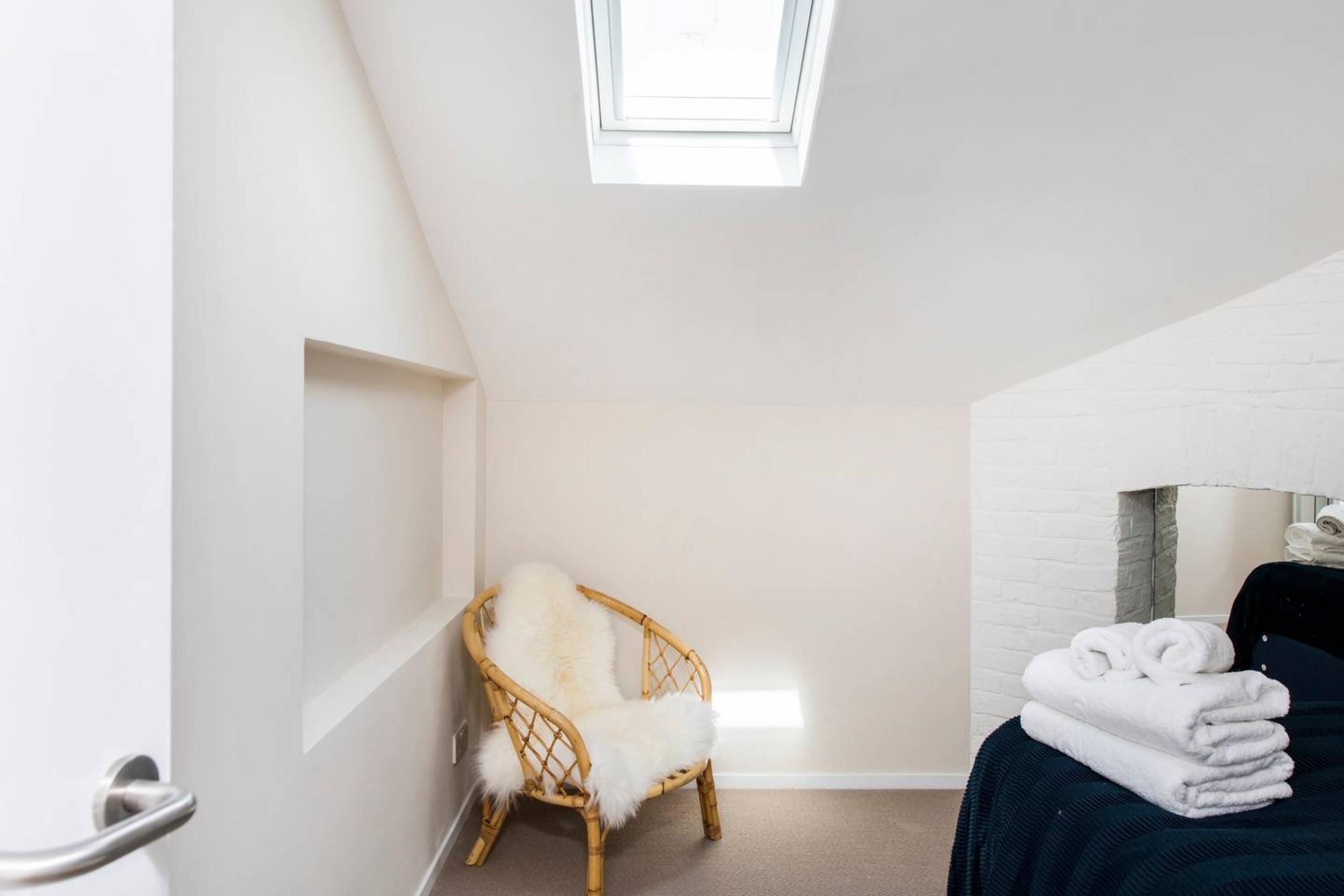 Ein kleiner Raum mit weißen Wänden und schrägen Decken, mit einem Oberlicht. Ein Korbstuhl mit flauschigem weißen Kissen steht an der Wand, und ein Stapel gefalteter weißer Handtücher liegt auf einem dunkelbraunen Bett.