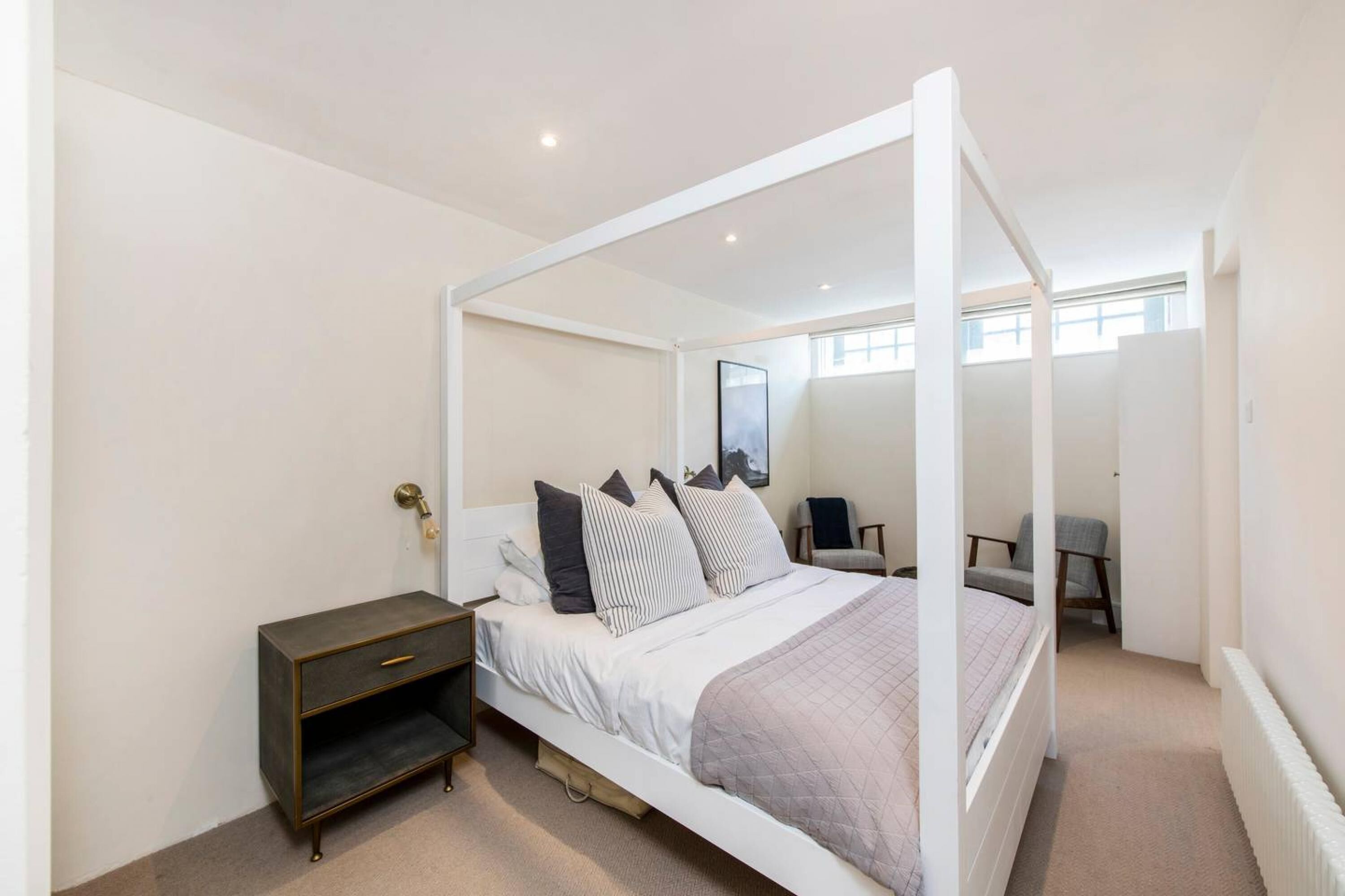 A four-poster bed with a white frame, a bedside table with a dark finish, and a chair in the background.